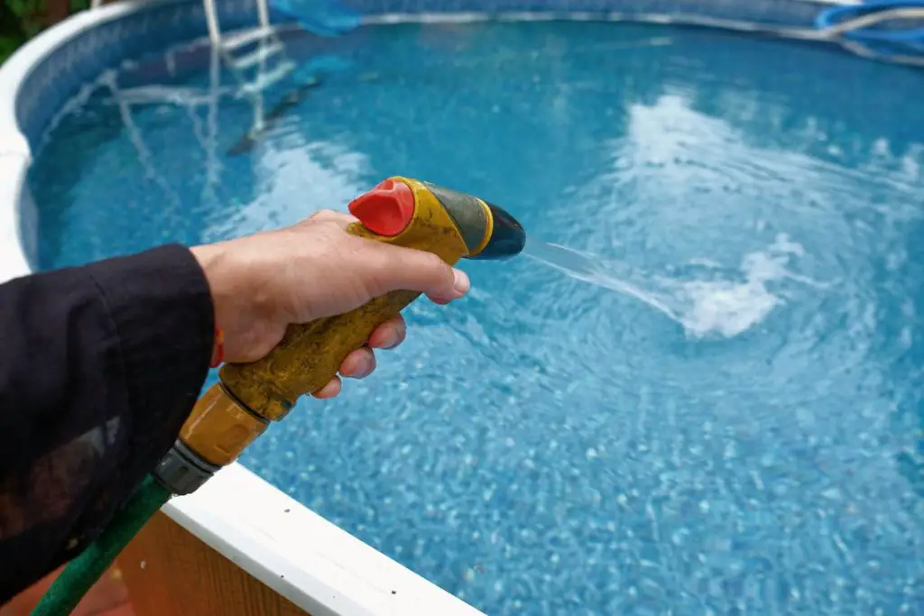 A hand with a garden hose pouring water into above ground swimming pool