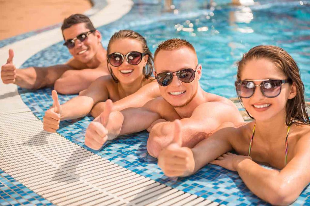 Beautiful young people having fun in swimming pool, smiling and showing thumbs up.